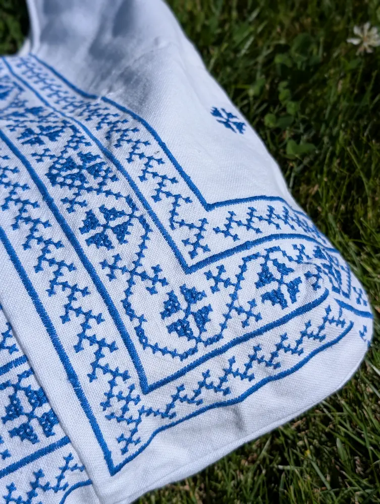 Detaliu geantă rustică din pânză albă cu flori albastre și motive geometrice, brodate în stil tradițional, din colecția de Sânziene
